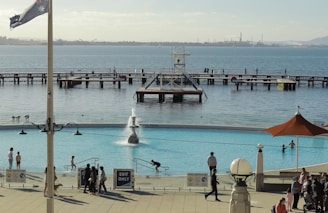 A waterfront scene features a large swimming pool with several people enjoying the water. The pool is surrounded by a terrace with visitors walking and relaxing. Beyond the pool, there is a body of water with piers and individuals strolling along. An Australian flag flies on a pole near the foreground, and the backdrop includes an industrial-looking shoreline.