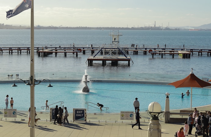 A waterfront scene features a large swimming pool with several people enjoying the water. The pool is surrounded by a terrace with visitors walking and relaxing. Beyond the pool, there is a body of water with piers and individuals strolling along. An Australian flag flies on a pole near the foreground, and the backdrop includes an industrial-looking shoreline.