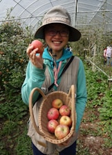 A person wearing glasses and a wide-brimmed hat is smiling and holding up an apple in one hand. The person is standing in a lush garden or orchard filled with greenery and apple trees. They are also holding a wicker basket containing several apples.