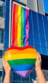 A rainbow flag is prominently displayed on a building. In the foreground, two hands are holding up a heart-shaped object with rainbow stripes matching the flag.