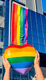 A rainbow flag is prominently displayed on a building. In the foreground, two hands are holding up a heart-shaped object with rainbow stripes matching the flag.