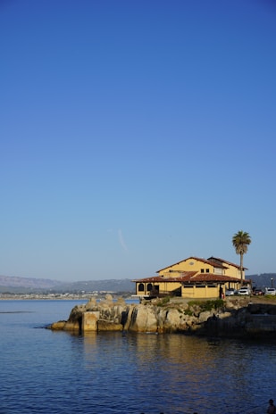 A large house with yellow walls and a red-tiled roof is situated on a rocky shoreline, adjacent to calm blue water. A tall palm tree stands nearby, and a few people and vehicles are visible near the house. The sky is clear and blue, with a mountainous landscape in the distance across the water.
