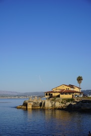 A large house with yellow walls and a red-tiled roof is situated on a rocky shoreline, adjacent to calm blue water. A tall palm tree stands nearby, and a few people and vehicles are visible near the house. The sky is clear and blue, with a mountainous landscape in the distance across the water.