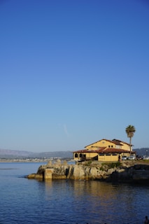 A large house with yellow walls and a red-tiled roof is situated on a rocky shoreline, adjacent to calm blue water. A tall palm tree stands nearby, and a few people and vehicles are visible near the house. The sky is clear and blue, with a mountainous landscape in the distance across the water.