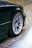 A close-up view of a car's silver alloy wheel and tire on a dark-colored vehicle, parked on a cobblestone street with some greenery growing in between the stones.