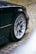 A close-up view of a car's silver alloy wheel and tire on a dark-colored vehicle, parked on a cobblestone street with some greenery growing in between the stones.