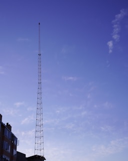 A tall, thin antenna tower rises against a clear blue sky with scattered clouds. The base of the tower is attached to the roof of a building, which is partially visible in the lower left corner.