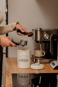 A person prepares coffee by scooping ground coffee with a measuring spoon into a portafilter. The setting includes a coffee machine and a ceramic mug on a wooden countertop. A tin labeled 'coffee' is used for storing the ground coffee.