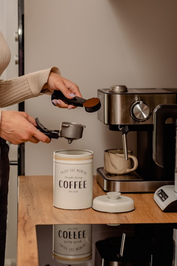 A person prepares coffee by scooping ground coffee with a measuring spoon into a portafilter. The setting includes a coffee machine and a ceramic mug on a wooden countertop. A tin labeled 'coffee' is used for storing the ground coffee.