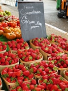 Baskets filled with fresh strawberries are on display at a market stall. Next to the strawberries, there are various types of tomatoes, including red and yellow ones. A black chalkboard sign with writing on it is situated in the middle of the display. The setting appears to be an outdoor market with asphalt ground and other market stalls visible in the background.