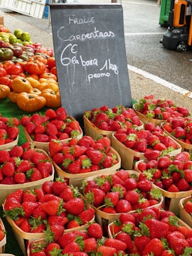 Baskets filled with fresh strawberries are on display at a market stall. Next to the strawberries, there are various types of tomatoes, including red and yellow ones. A black chalkboard sign with writing on it is situated in the middle of the display. The setting appears to be an outdoor market with asphalt ground and other market stalls visible in the background.