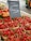 Baskets filled with fresh strawberries are on display at a market stall. Next to the strawberries, there are various types of tomatoes, including red and yellow ones. A black chalkboard sign with writing on it is situated in the middle of the display. The setting appears to be an outdoor market with asphalt ground and other market stalls visible in the background.