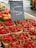 Baskets filled with fresh strawberries are on display at a market stall. Next to the strawberries, there are various types of tomatoes, including red and yellow ones. A black chalkboard sign with writing on it is situated in the middle of the display. The setting appears to be an outdoor market with asphalt ground and other market stalls visible in the background.