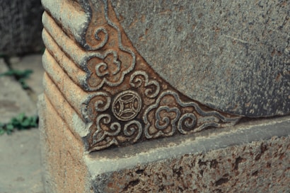 An ornate stone structure with elaborate carvings featuring swirling patterns and floral motifs. The stone texture appears weathered, and there is a subtle blue-gray color integrated with the natural stone hues.