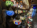 A collection of colorful mosaic glass lamps with intricate patterns is displayed on multiple shelves. The lamps feature vibrant hues and ornate designs, creating an inviting and exotic ambiance in what appears to be a shop or market setting.