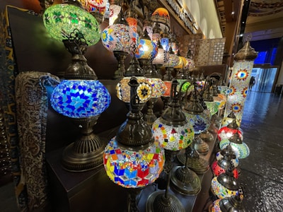 A collection of colorful mosaic glass lamps with intricate patterns is displayed on multiple shelves. The lamps feature vibrant hues and ornate designs, creating an inviting and exotic ambiance in what appears to be a shop or market setting.