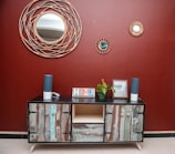 A rustic wooden sideboard with an eclectic design stands against a deep red wall. Above it, three decorative round mirrors with woven frames add an artistic touch. The sideboard is adorned with two tall blue speakers, a potted green plant, a picture frame, and a colorful 'HOME' sign. The floor is tiled in a neutral beige color, complementing the decor.