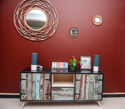 A rustic wooden sideboard with an eclectic design stands against a deep red wall. Above it, three decorative round mirrors with woven frames add an artistic touch. The sideboard is adorned with two tall blue speakers, a potted green plant, a picture frame, and a colorful 'HOME' sign. The floor is tiled in a neutral beige color, complementing the decor.