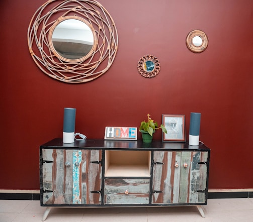 A rustic wooden sideboard with an eclectic design stands against a deep red wall. Above it, three decorative round mirrors with woven frames add an artistic touch. The sideboard is adorned with two tall blue speakers, a potted green plant, a picture frame, and a colorful 'HOME' sign. The floor is tiled in a neutral beige color, complementing the decor.