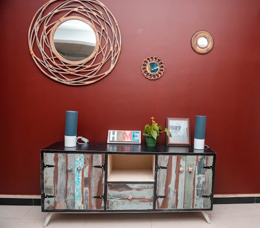 A rustic wooden sideboard with an eclectic design stands against a deep red wall. Above it, three decorative round mirrors with woven frames add an artistic touch. The sideboard is adorned with two tall blue speakers, a potted green plant, a picture frame, and a colorful 'HOME' sign. The floor is tiled in a neutral beige color, complementing the decor.