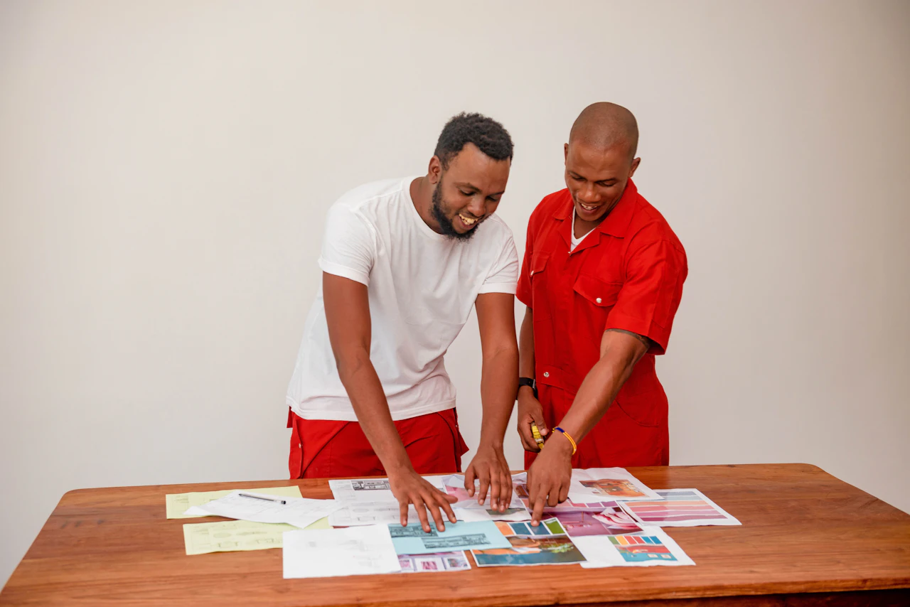 Two smiling decorators, one in a red boiler suit, choose paint colours from swatches spread on a wooden table.