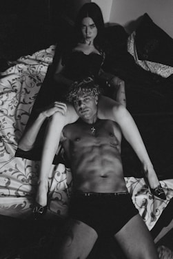 A black and white photograph displays a shirtless man lying on a bed, with a woman sitting behind him in a dark outfit. The setting is intimate, with patterned bedding and dim lighting.