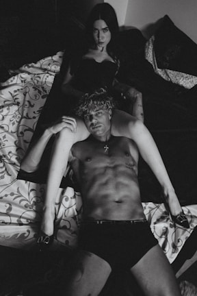 A black and white photograph displays a shirtless man lying on a bed, with a woman sitting behind him in a dark outfit. The setting is intimate, with patterned bedding and dim lighting.