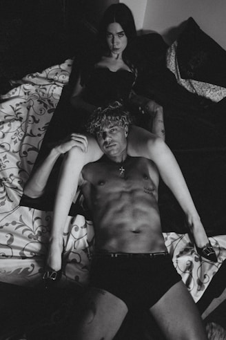 A black and white photograph displays a shirtless man lying on a bed, with a woman sitting behind him in a dark outfit. The setting is intimate, with patterned bedding and dim lighting.