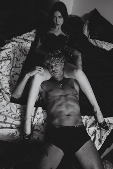 A black and white photograph displays a shirtless man lying on a bed, with a woman sitting behind him in a dark outfit. The setting is intimate, with patterned bedding and dim lighting.
