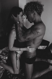 A black and white photograph of two people in an intimate embrace, with tattoos visible on one person's arms. They are kneeling on a bed, creating a close and personal atmosphere.