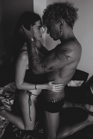A black and white photograph of two people in an intimate embrace, with tattoos visible on one person's arms. They are kneeling on a bed, creating a close and personal atmosphere.