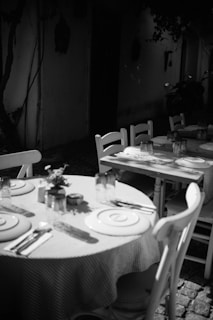 An outdoor dining area with a rustic ambiance, featuring round tables covered with checkered tablecloths, set with plates, cutlery, and glassware. The scene is in black and white, creating a nostalgic and timeless feeling. Shadows from chairs and other elements suggest natural lighting.