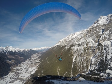A paraglider is soaring through the sky with a vast mountain landscape below. Snow-capped peaks and valleys extend into the distance with a village nestled in the valley below. The sky is clear, allowing for an expansive view of the white and green terrain.