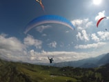 Several paragliders are soaring high above a lush, green mountainous landscape. The sky is bright and filled with scattered clouds, and the sun is shining brightly, casting a warm glow. The paragliders have canopies of various colors, including blue and red, adding vivid contrast to the natural scene.