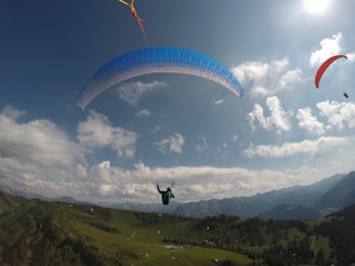 Several paragliders are soaring high above a lush, green mountainous landscape. The sky is bright and filled with scattered clouds, and the sun is shining brightly, casting a warm glow. The paragliders have canopies of various colors, including blue and red, adding vivid contrast to the natural scene.
