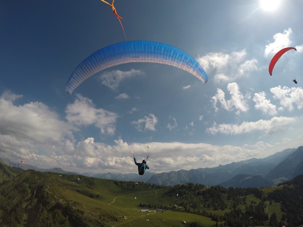 Several paragliders are soaring high above a lush, green mountainous landscape. The sky is bright and filled with scattered clouds, and the sun is shining brightly, casting a warm glow. The paragliders have canopies of various colors, including blue and red, adding vivid contrast to the natural scene.