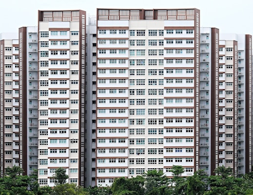 A series of high-rise apartment buildings with a symmetrical design and predominantly white and brown colors. The buildings feature numerous windows and balconies. In the foreground, lush green trees add a touch of nature to the urban landscape.