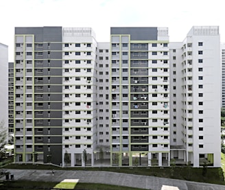 A modern, multi-story residential apartment building with a symmetrical facade, featuring alternating rows of windows and balconies. The structure is primarily composed of two colors, green and white, with a clear division of levels visible. There's a pathway in the foreground and some sparse trees.