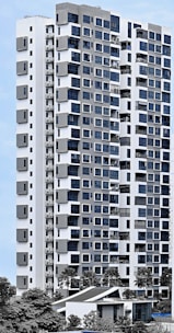 A modern high-rise residential building with a sleek facade consisting of a mix of large windows and geometric patterns. The structure is predominantly white with grey accents, surrounded by greenery and palm trees. The sky is clear, providing a clean backdrop for the building.