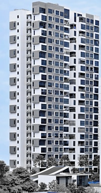 A modern high-rise residential building with a sleek facade consisting of a mix of large windows and geometric patterns. The structure is predominantly white with grey accents, surrounded by greenery and palm trees. The sky is clear, providing a clean backdrop for the building.