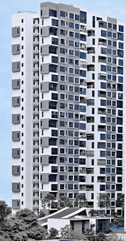 A modern high-rise residential building with a sleek facade consisting of a mix of large windows and geometric patterns. The structure is predominantly white with grey accents, surrounded by greenery and palm trees. The sky is clear, providing a clean backdrop for the building.