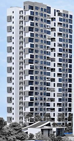 A modern high-rise residential building with a sleek facade consisting of a mix of large windows and geometric patterns. The structure is predominantly white with grey accents, surrounded by greenery and palm trees. The sky is clear, providing a clean backdrop for the building.