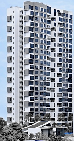 A modern high-rise residential building with a sleek facade consisting of a mix of large windows and geometric patterns. The structure is predominantly white with grey accents, surrounded by greenery and palm trees. The sky is clear, providing a clean backdrop for the building.