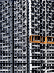 Tall residential buildings with a modern architectural design, featuring numerous windows arranged in an orderly, repetitive pattern. Some sections are highlighted with orange accents.