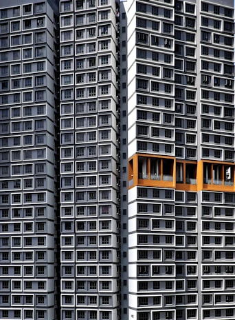 Tall residential buildings with a modern architectural design, featuring numerous windows arranged in an orderly, repetitive pattern. Some sections are highlighted with orange accents.