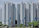 High-rise residential buildings with a modern design featuring a combination of white and light blue colors. The buildings are situated in an urban area with lush green trees in the foreground.
