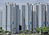 High-rise residential buildings with a modern design featuring a combination of white and light blue colors. The buildings are situated in an urban area with lush green trees in the foreground.