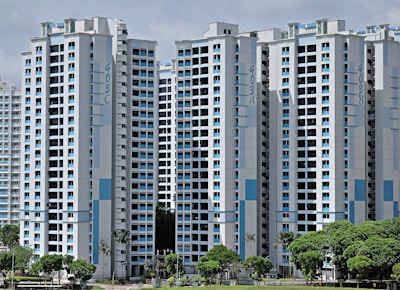 High-rise residential buildings with a modern design featuring a combination of white and light blue colors. The buildings are situated in an urban area with lush green trees in the foreground.