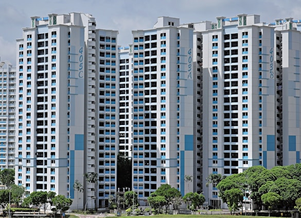High-rise residential buildings with a modern design featuring a combination of white and light blue colors. The buildings are situated in an urban area with lush green trees in the foreground.