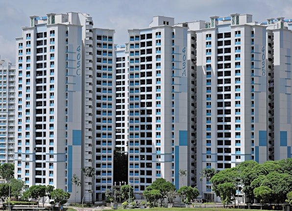 High-rise residential buildings with a modern design featuring a combination of white and light blue colors. The buildings are situated in an urban area with lush green trees in the foreground.